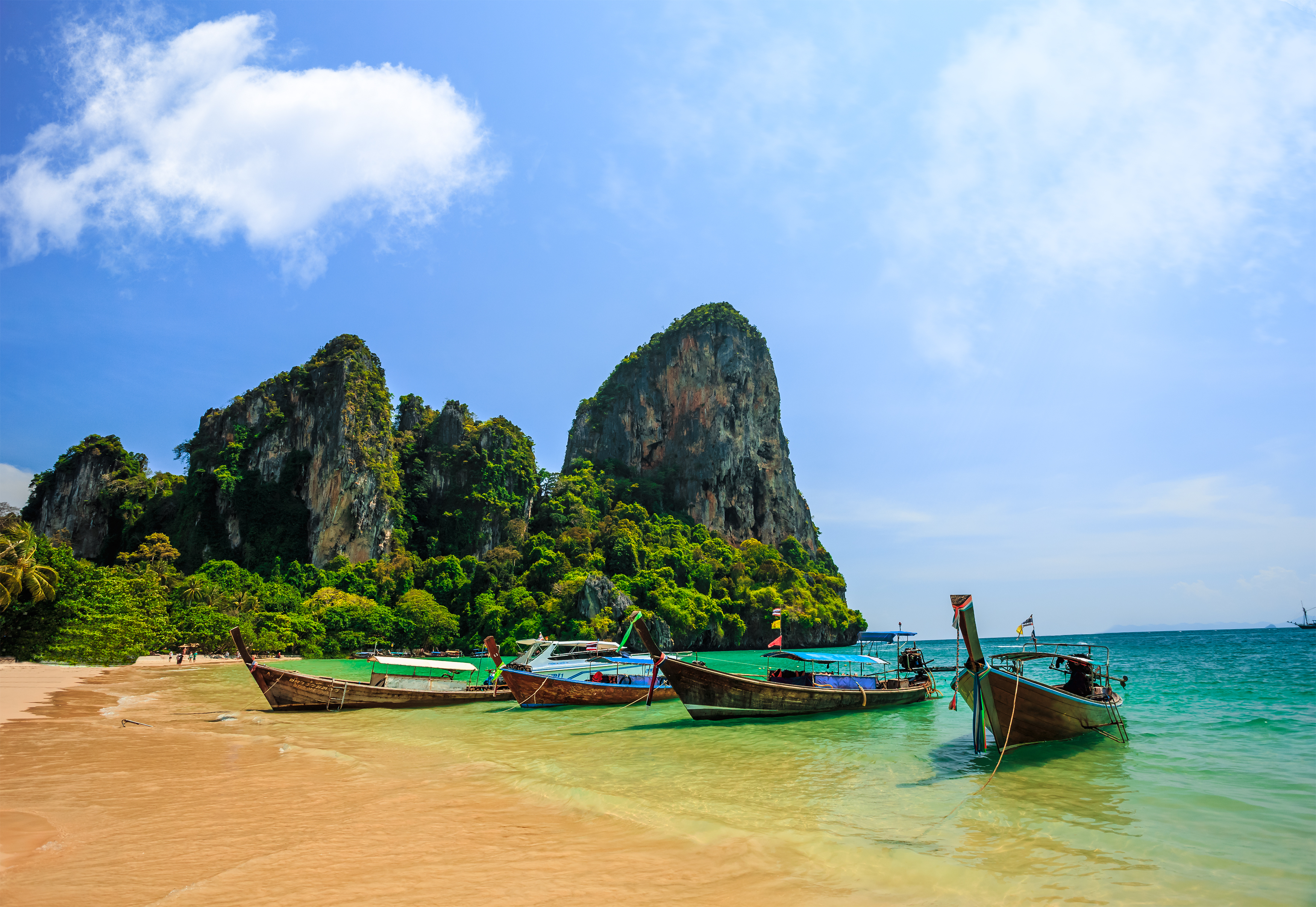 Los acantilados adornados con musgo y hierba se elevan de fondo mientras el mar choca contra la orilla, todo mientras los barcos de pesca flotan en el mar. Estar en la playa de Ao Nang es una experiencia tailandesa única