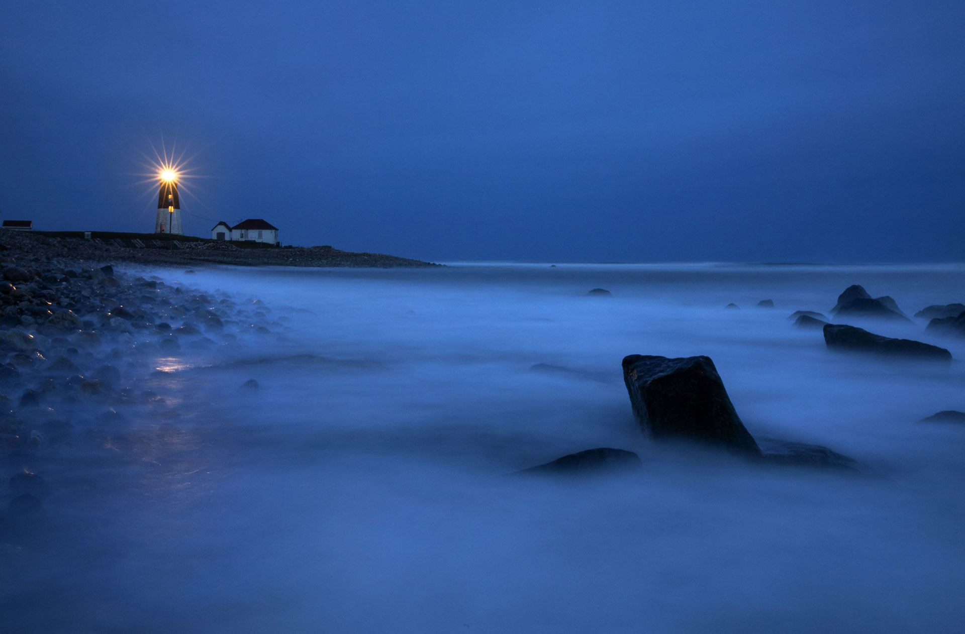 Las olas bañan las rocas cuando un faro de luz brilla a través de la costa rocosa desde el faro de Point Judith en Narragansett, Rhode Island, EEUU, el sábado 25 de abril de 2020. (Foto AP / David Goldman)